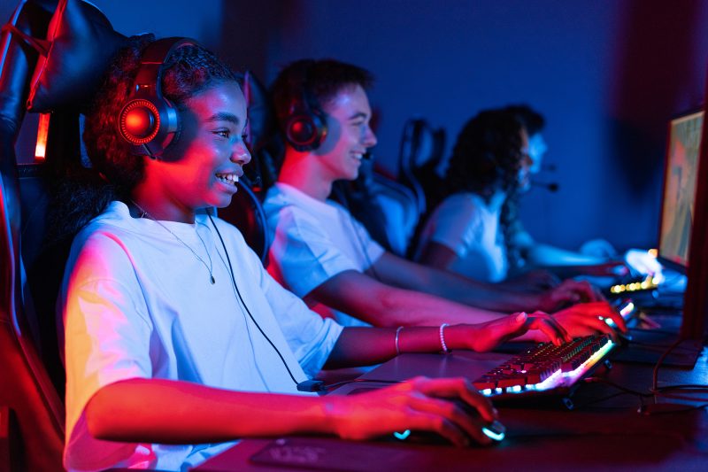 Group of multiracial teens in headsets playing video games in video game club with blue and red illumination. Keyboard and mouse with illumination